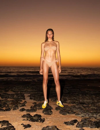 Model Angelina Kendall in a striped bikini and yellow sneakers standing on a rocky shore at sunset for Pop Magazine.
