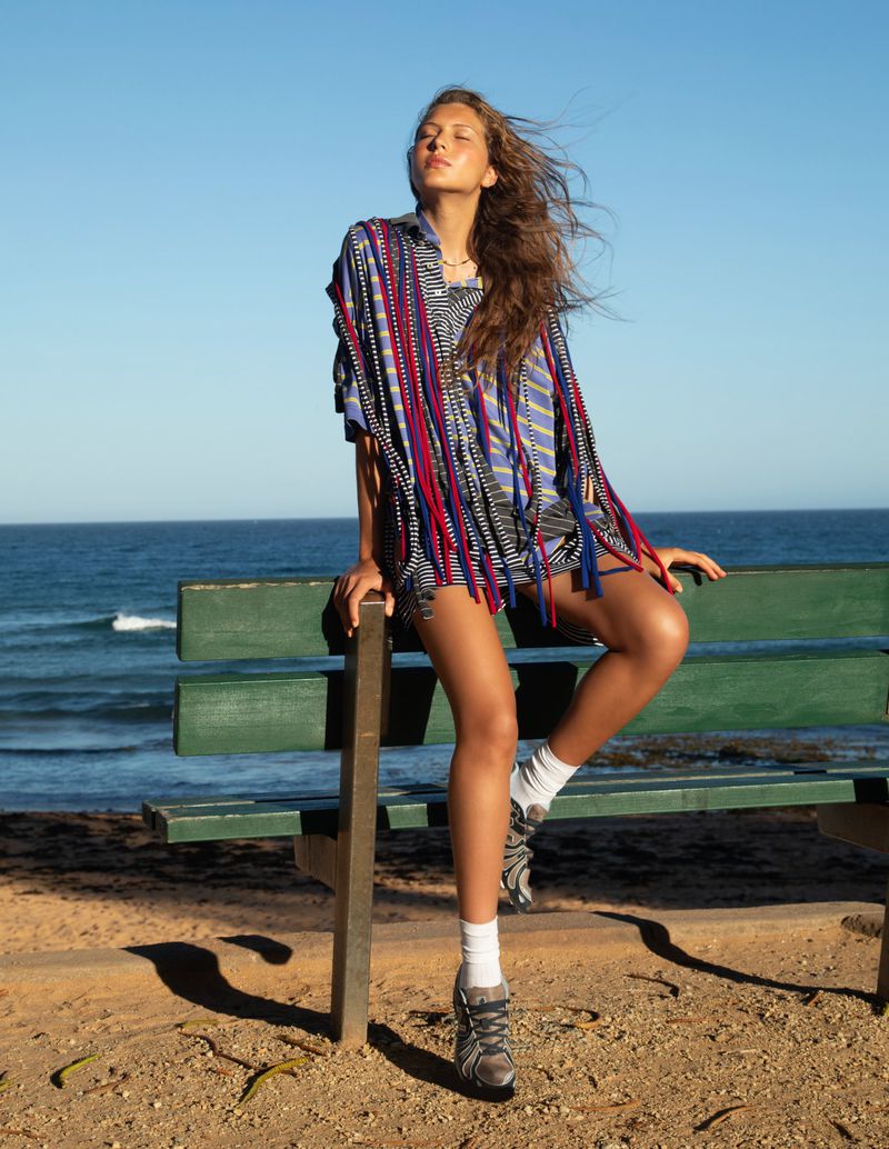 Model Angelina Kendall seated on a green park bench by the ocean wearing a striped shirt with colorful fringe.
