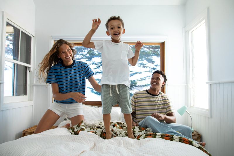 A young boy jumping on a bed with a smiling woman and man watching in a sunlit bedroom