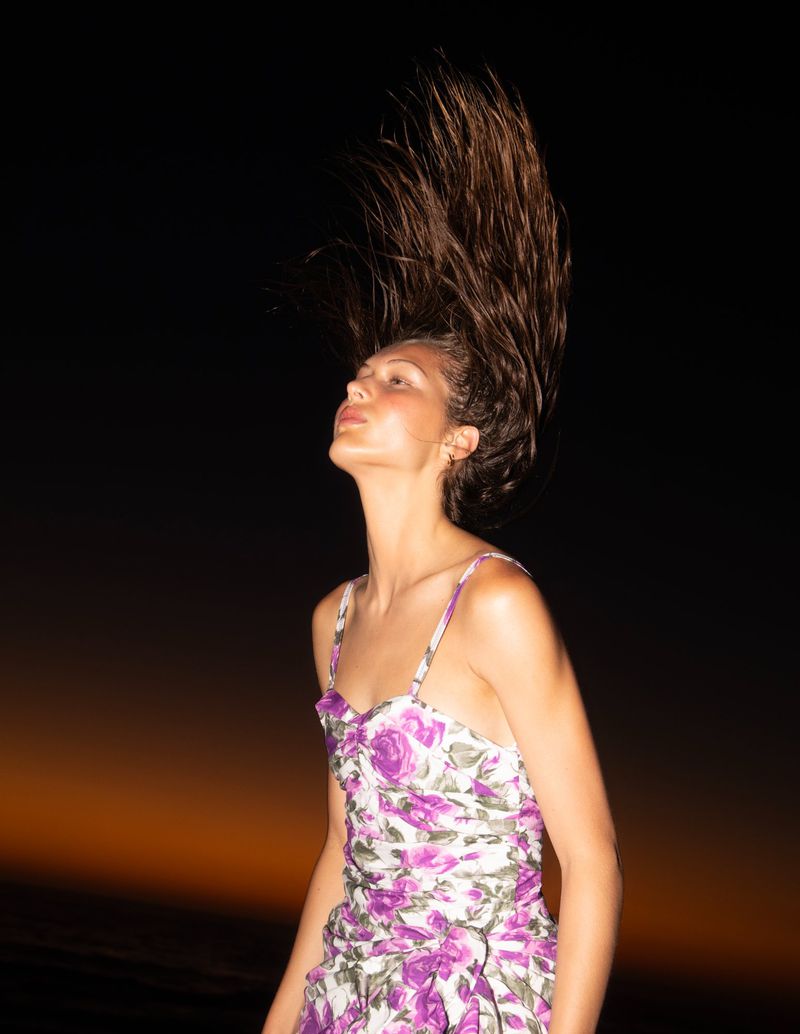 Woman with long brown hair tosses her head back, wearing a floral dress in a dark outdoor setting at dusk. Fashion shot, beauty.