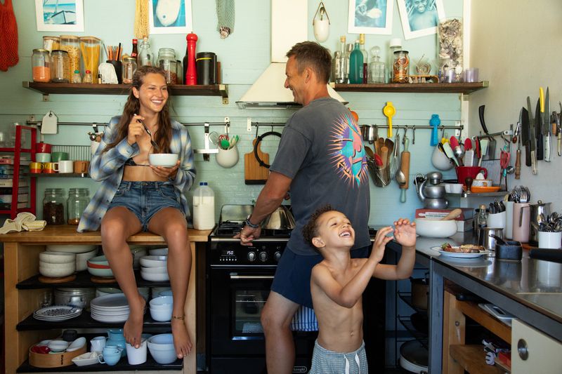 Young woman in plaid shirt seated on kitchen counter with man and child in kitchen for Pop Magazine