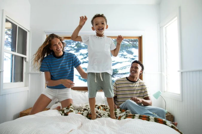 A young boy jumping on a bed with a smiling woman and man watching in a sunlit bedroom