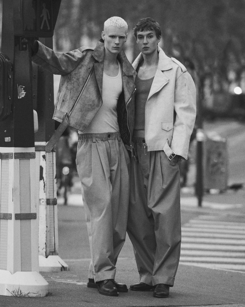 Two male models standing side by side on a city street wearing layered jackets and pleated trousers in a black and white editorial photo.