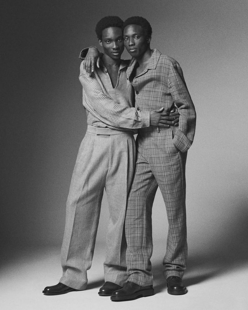 Two black men posing together wearing patterned long-sleeve shirts and trousers against a grey seamless studio background.