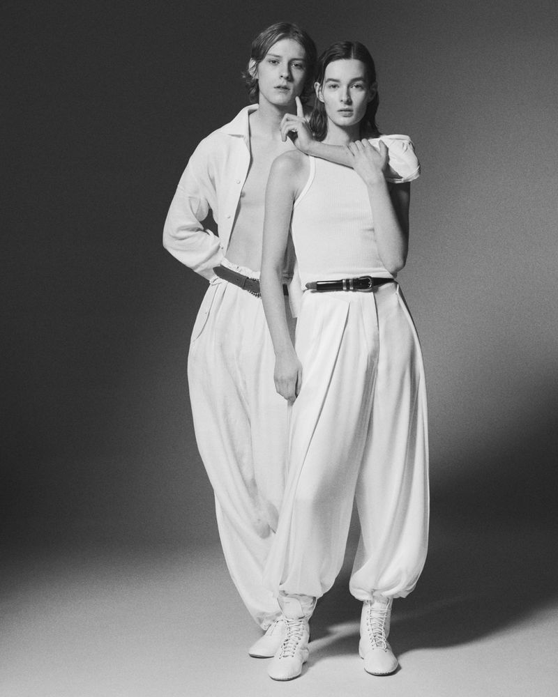 Two models wearing white trousers and matching white tops posing together in a studio setting against a neutral background.
