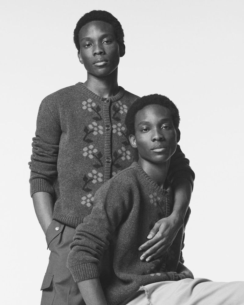 Monochrome portrait of two Black male fashion models in matching cardigans, one standing behind the other in a light-colored studio.