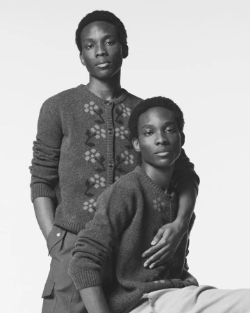 Monochrome portrait of two Black male fashion models in matching cardigans, one standing behind the other in a light-colored studio.