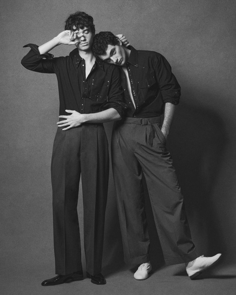 Two men posing together in dark shirts and trousers against a neutral textured studio background, photographed by Laurent Humbert.