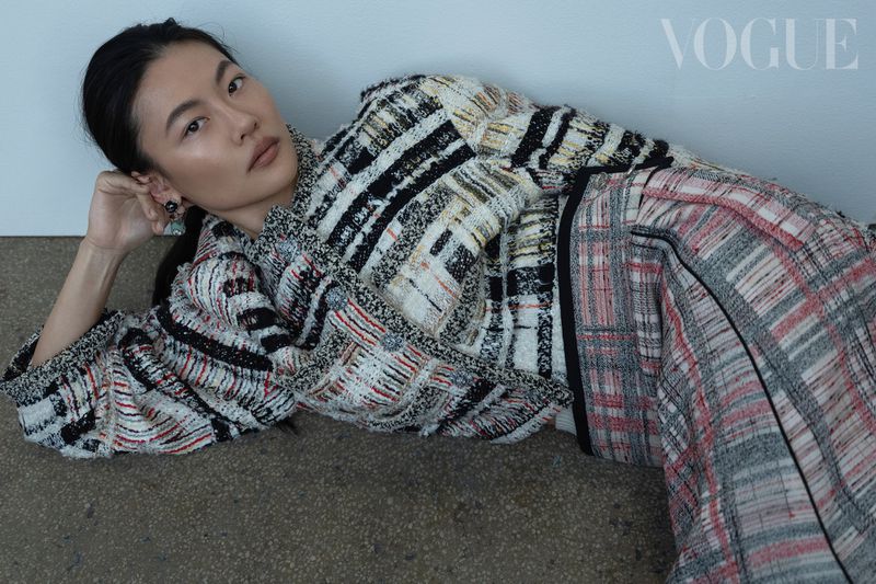 East Asian woman lying on floor wearing patterned tweed jacket and skirt against plain wall