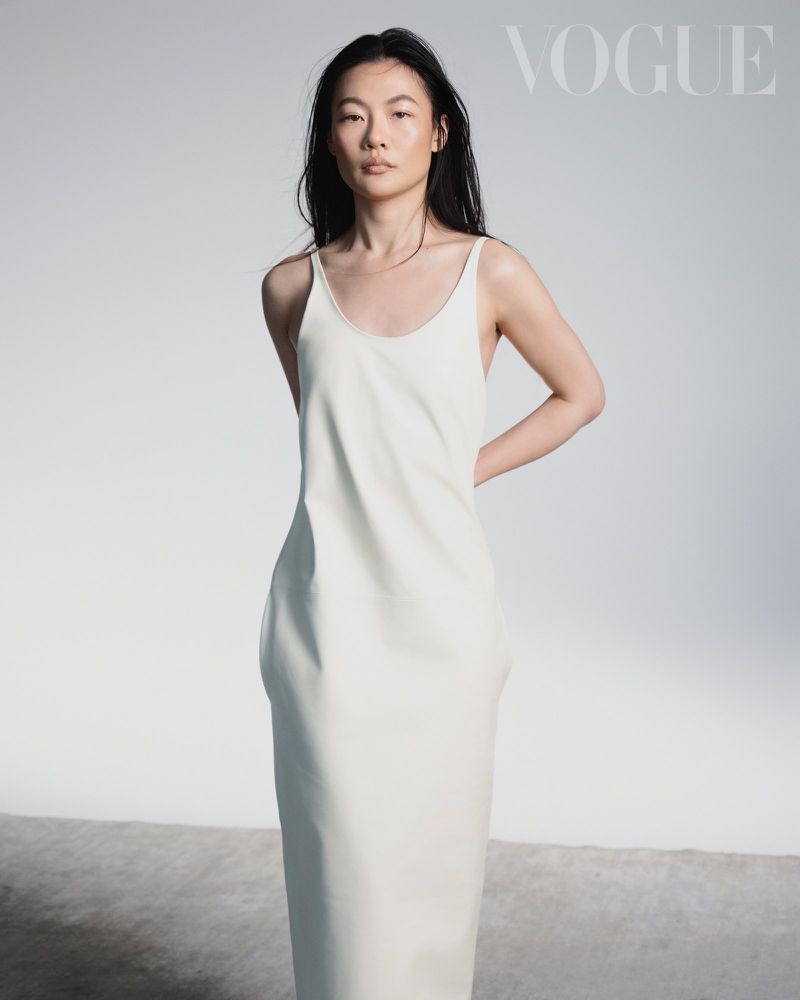 Woman with long dark hair wearing a sleeveless cream slip dress against a plain white studio background with Vogue text.