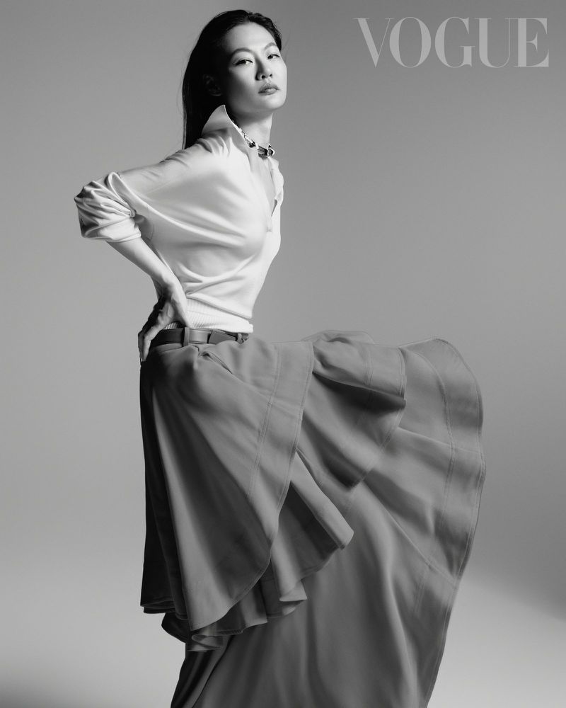 Model in white collared shirt and layered skirt posing against a seamless grey studio backdrop for Vogue Taiwan