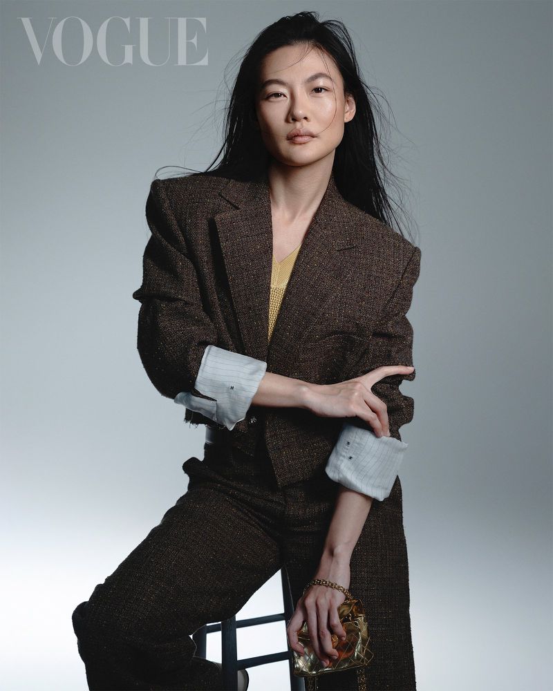 Asian woman wearing brown tweed jacket and trousers holding small gold handbag against plain grey studio backdrop
