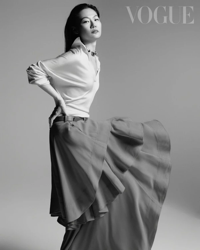 Model in white collared shirt and layered skirt posing against a seamless grey studio backdrop for Vogue Taiwan