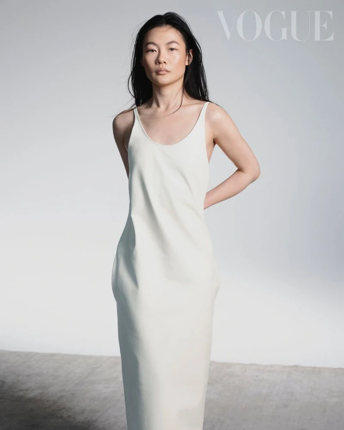 Woman with long dark hair wearing a sleeveless cream slip dress against a plain white studio background with Vogue text.