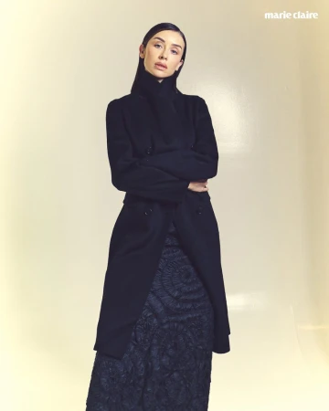 Claudia Bouza wearing a long black coat and textured skirt against a neutral studio background