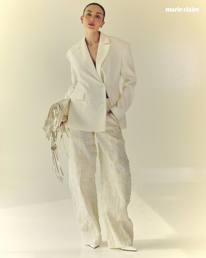 A stylish adult woman in a white double-breasted blazer and textured pants poses in a fashion editorial, holding a shimmering purse.