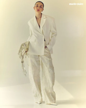 Model in white blazer and textured trousers holding a metallic fringed handbag against a neutral studio background.