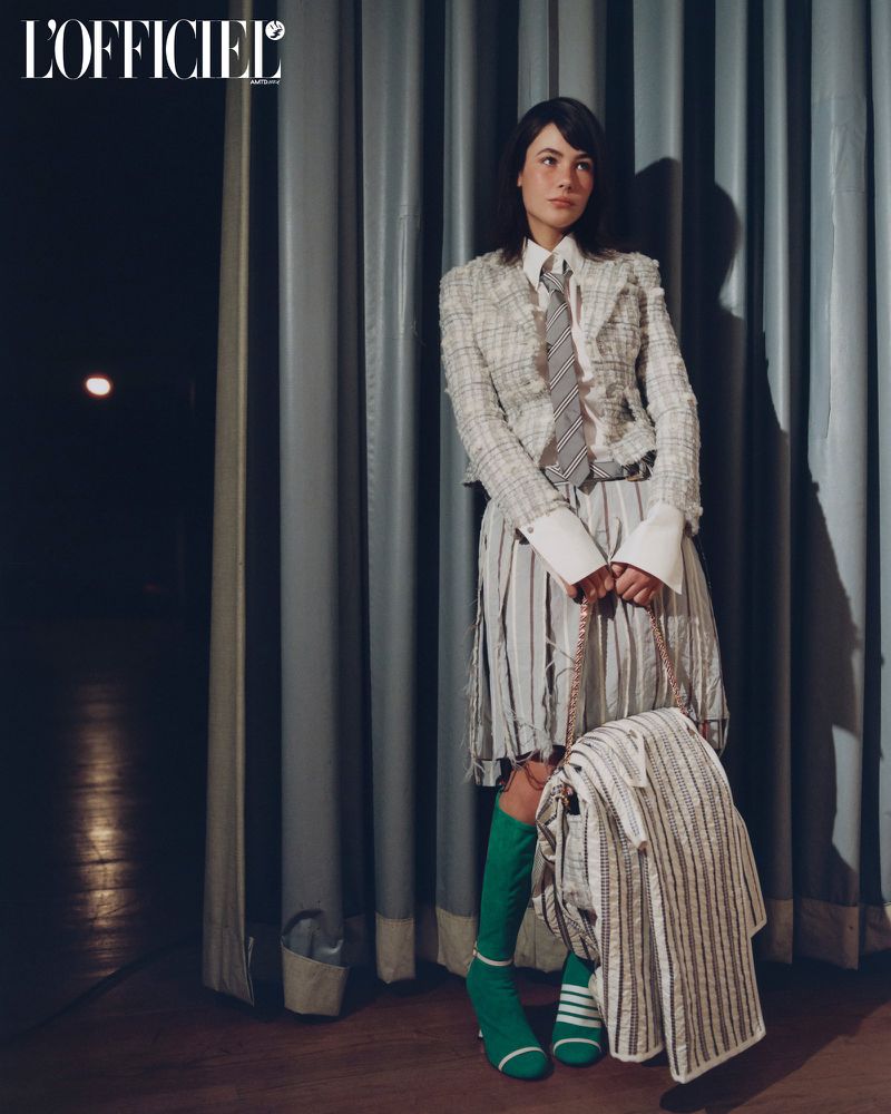 Young woman poses in a striped skirt and jacket in front of grey curtains in a fashion editorial shot with dark mood lighting.