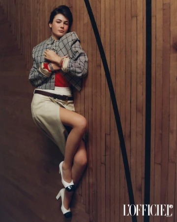 Grace Burns wearing a plaid jacket and skirt leaning against a wood panel wall for L'Officiel USA