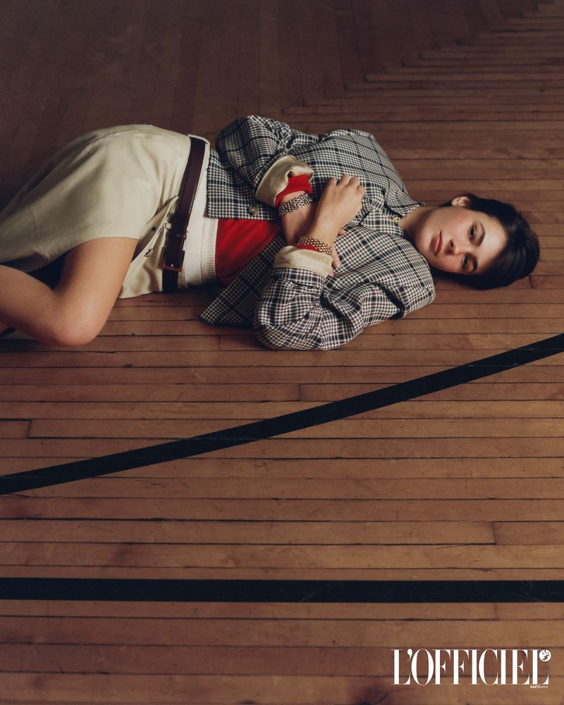 Model Grace Burns lying on a polished wooden floor wearing a plaid jacket and cream skirt for L'Officiel USA.