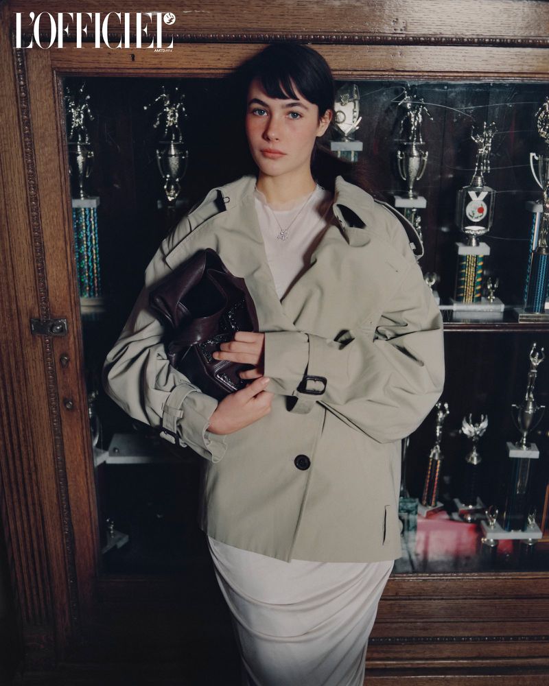 Model Grace Burns wearing a beige trench coat holding a dark handbag in front of a trophy cabinet for L'Officiel USA