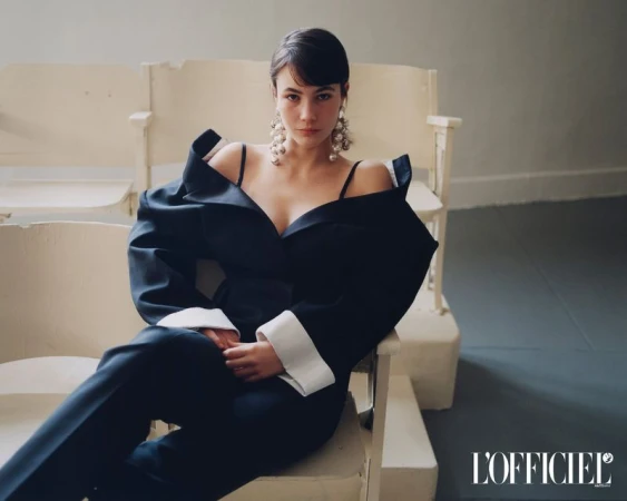 Grace Burns seated on a white chair wearing an off-the-shoulder dark suit jacket with statement pearl earrings.