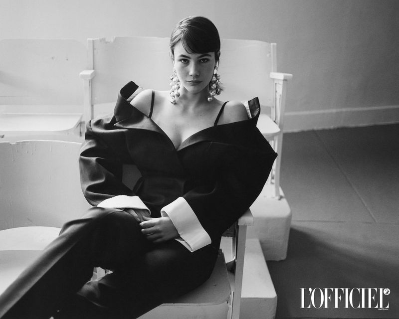 Model Grace Burns seated wearing a structured dark suit with white cuffs and large pearl earrings for L Officiel USA