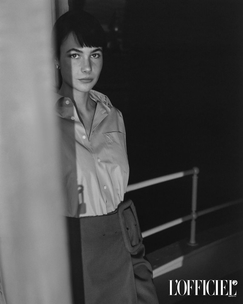 Black and white portrait of model Grace Burns wearing a buttoned shirt and skirt outdoors near a railing.