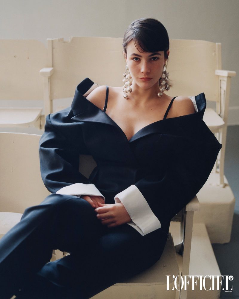 Model Grace Burns wearing a black structured blazer and large earrings seated on a chair for L'Officiel USA