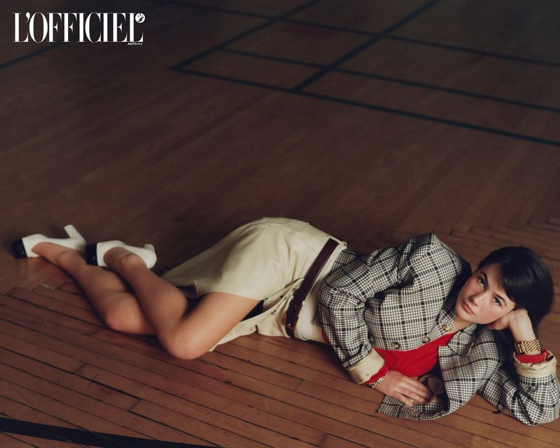 Model Grace Burns lying on a polished wooden floor wearing a plaid blazer, cream skirt, and white heels for L'Officiel USA.