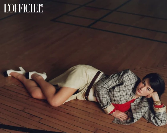 Model Grace Burns lying on a polished wooden floor wearing a plaid blazer, cream skirt, and white heels for L'Officiel USA.