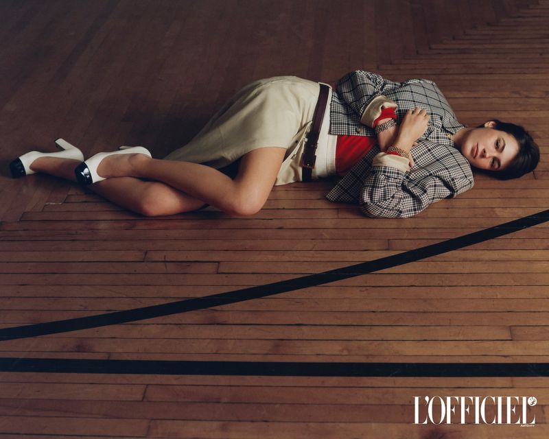 Model lying on a polished wooden gymnasium floor wearing a plaid blazer, cream skirt, and white heels for L'Officiel USA