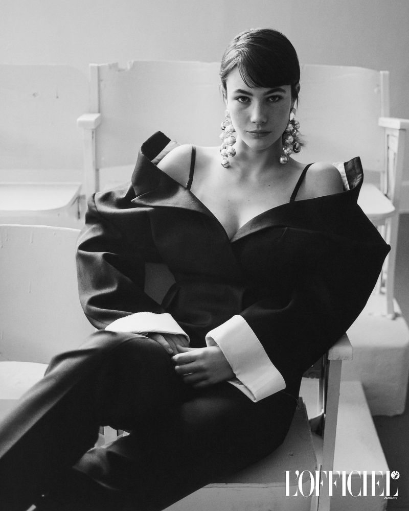 Model Grace Burns wearing a black structured blazer and statement pearl earrings, seated in a white wooden chair for L'Officiel USA.