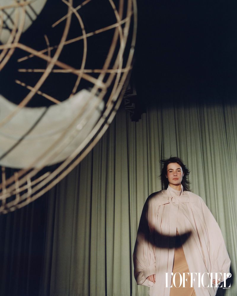 Model Grace Burns wearing a voluminous light-colored coat standing before green draped curtains with a foreground fan cage structure.