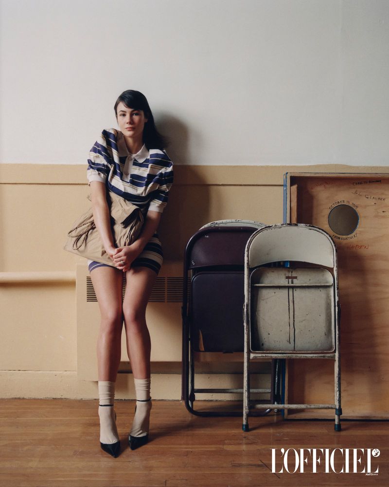 Model Grace Burns wearing a striped polo dress and beige handbag sitting against a wall with folding chairs in an L'Officiel USA feature