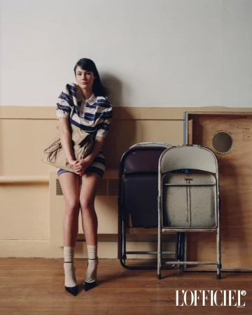 Model Grace Burns wearing a striped polo dress and beige handbag sitting against a wall with folding chairs in an L'Officiel USA feature