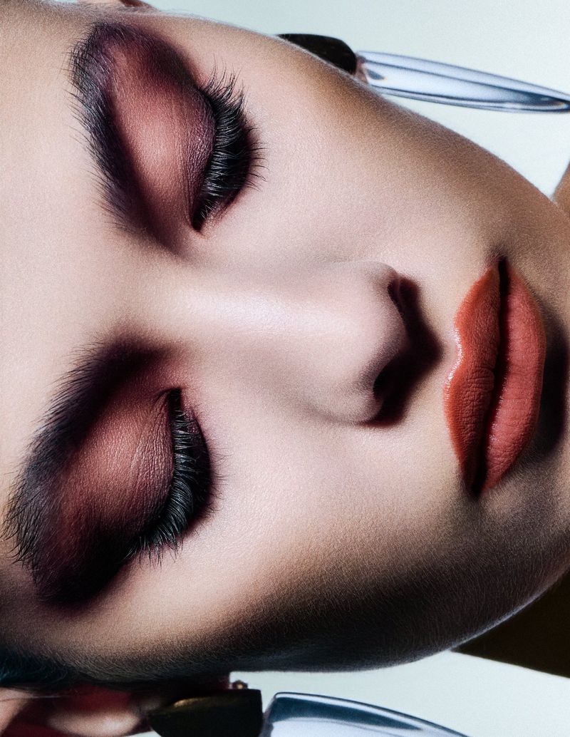 Close-up beauty portrait of a model with dramatic plum eyeshadow and sleek dark brows against a plain light background.