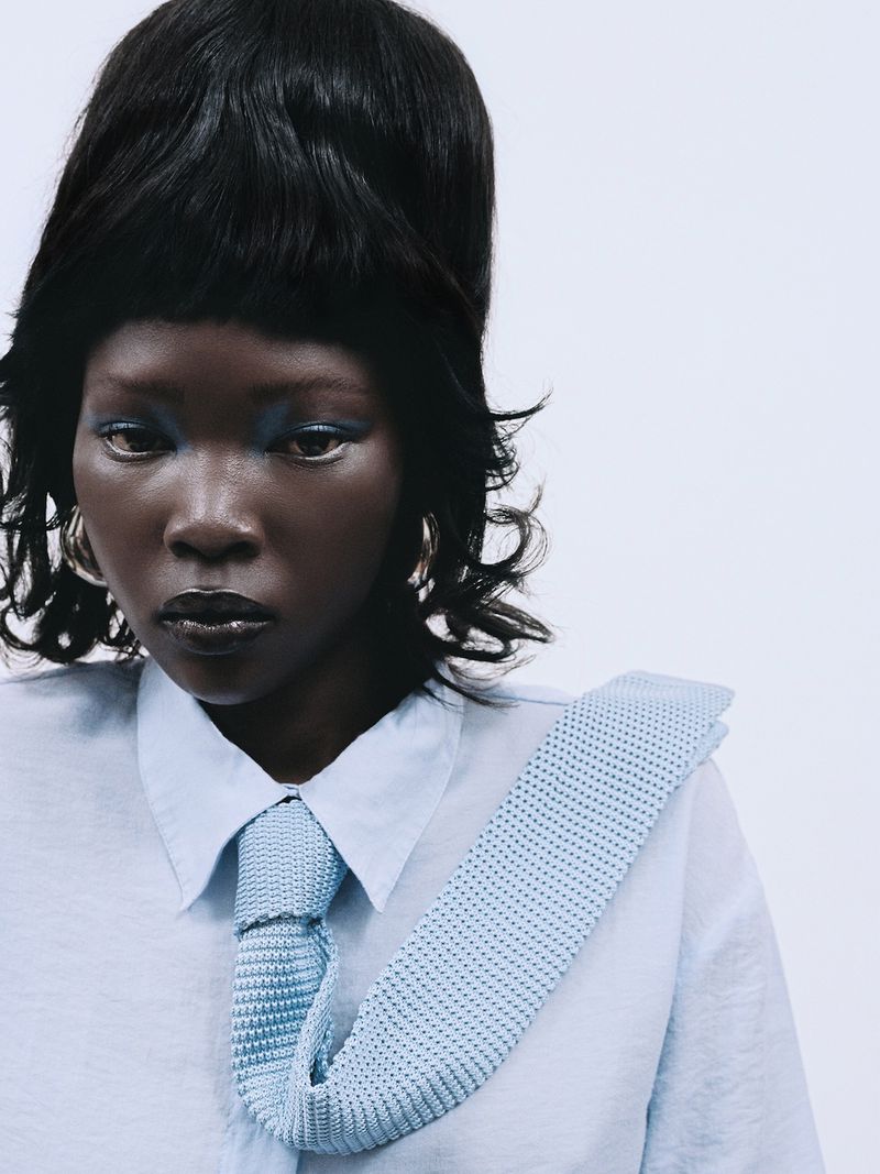 Model with voluminous dark hair and blue eyeshadow wearing a light blue shirt and tie against a plain white background