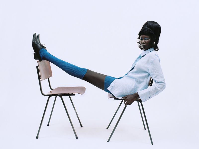 Black model with beehive hair resting legs on a second chair wearing blue socks and light blue shirt
