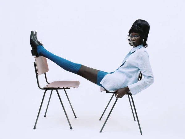 Black model with beehive hair resting legs on a second chair wearing blue socks and light blue shirt