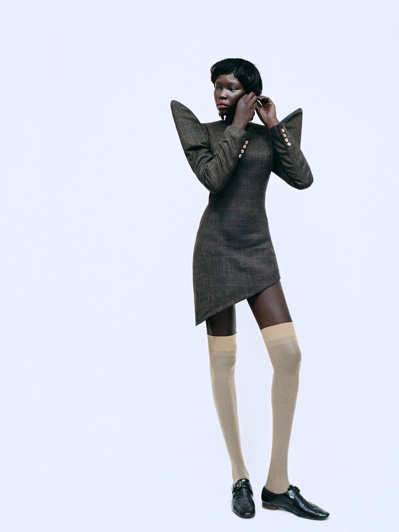 Model wearing a structured brown dress with exaggerated shoulder pads and knee-high stockings against a white background.