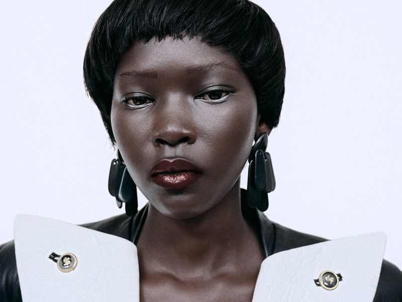 Close-up portrait of a black woman with short dark hair wearing statement earrings and a structured white-collared jacket against a white background.