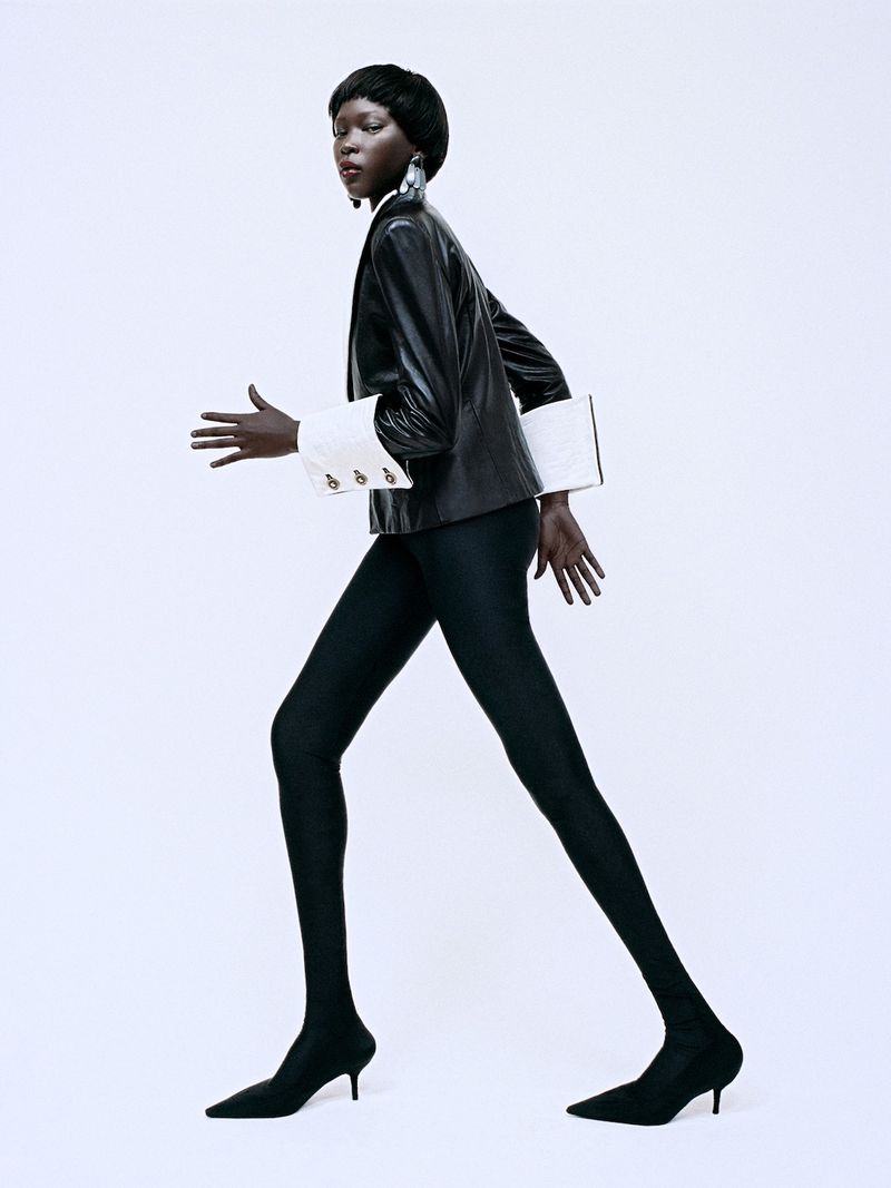An adult female model in a black jacket and leggings strides against a white backdrop, showcasing fashion apparel and minimalist style.