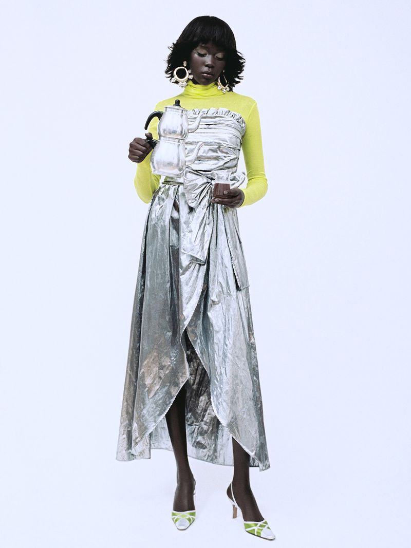 Model wearing a metallic silver wrap skirt and yellow turtleneck holding a silver kettle against a white background.