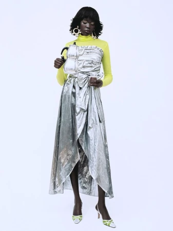Model wearing a metallic silver wrap skirt and yellow turtleneck holding a silver kettle against a white background.