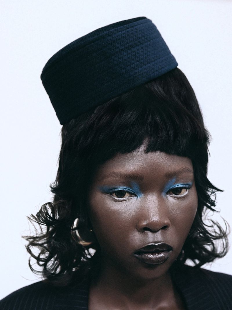 Portrait of a Black woman wearing a dark pillbox hat and bright blue eyeshadow against a plain white background