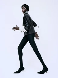 Model in black blazer with white cuffs and black leggings walking against a bright white background