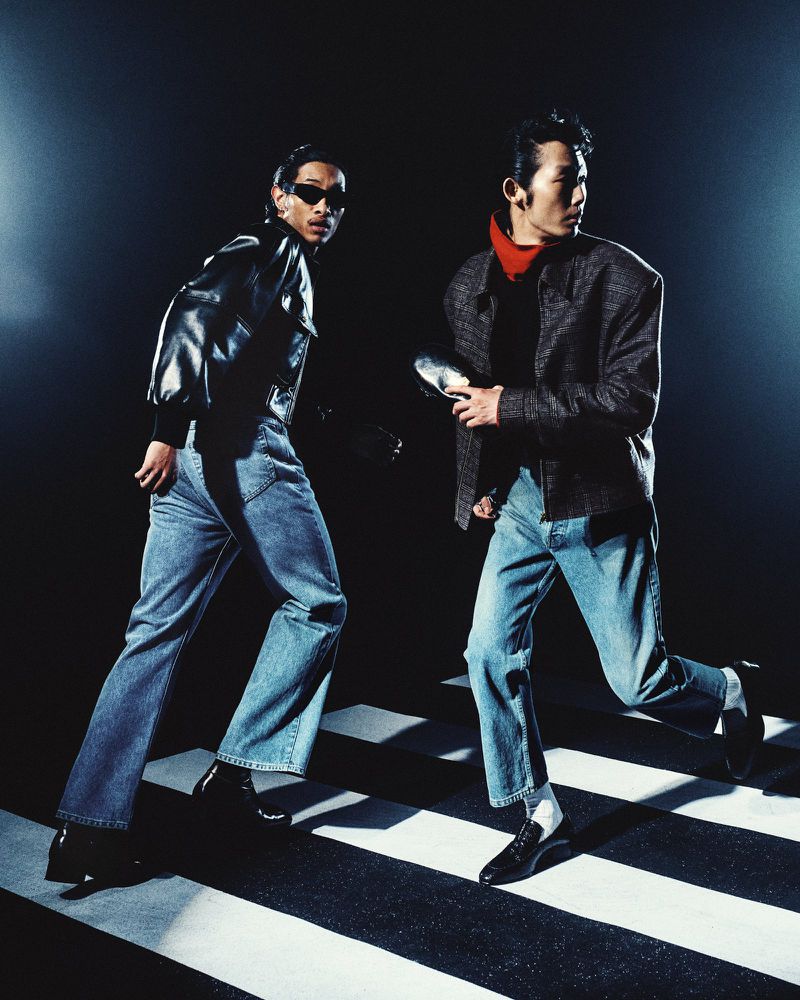 Two male models walking briskly on a striped studio floor wearing dark jackets and blue jeans for Willy Chavarria.