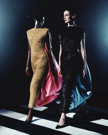 Two models wearing textured evening dresses with capes walking on a high contrast black and white striped floor under dramatic low-key lighting