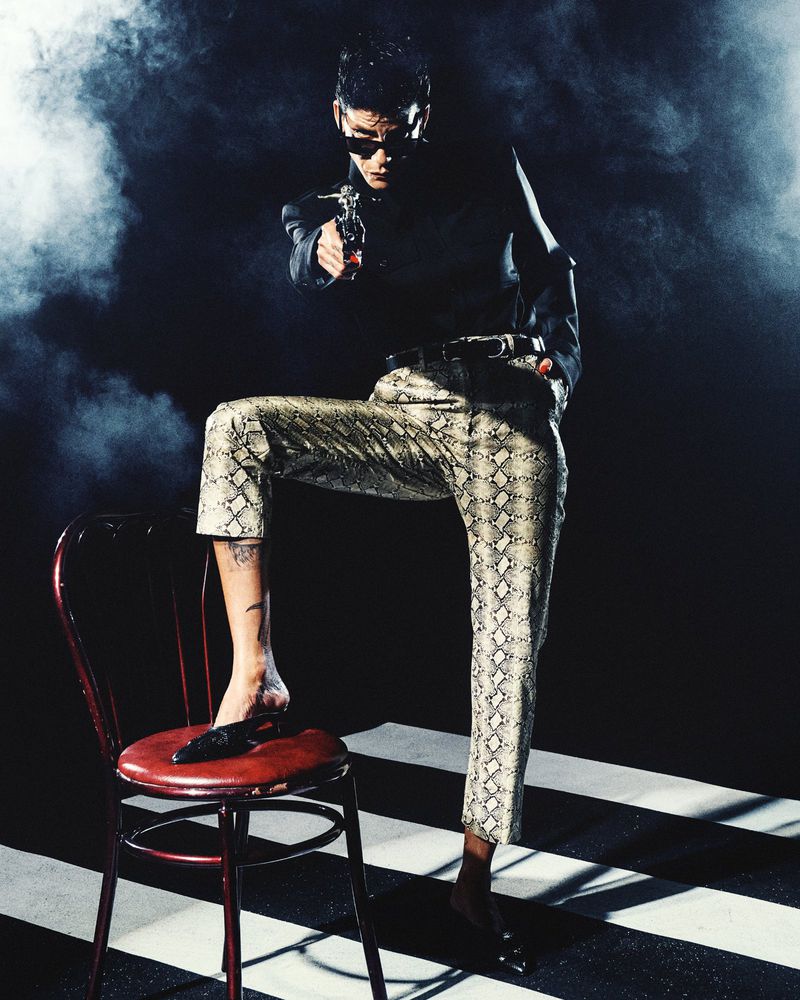 Man wearing sunglasses and snakeskin pants with one foot on a red chair aiming a small gun